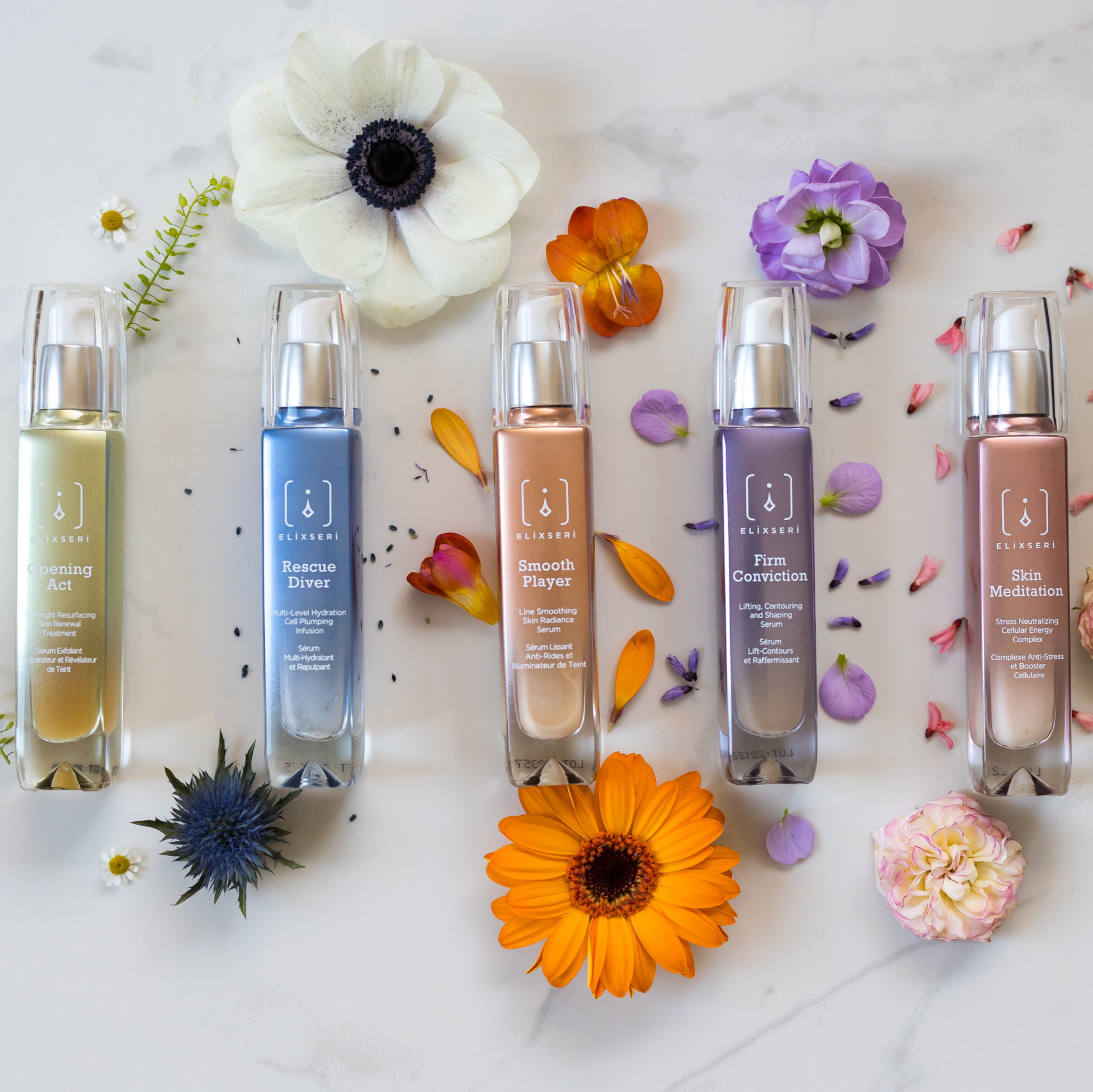 Colourful elixseri serum bottles lined up on a pale grey marble background with colourful spring flowers scattered around them