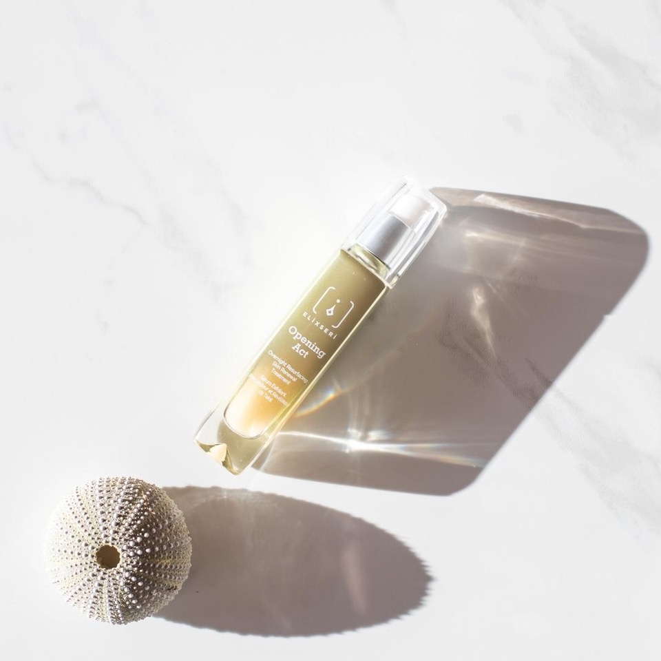 Elixseri's Opening Act serum pictured next to a white shell in summer sun. 