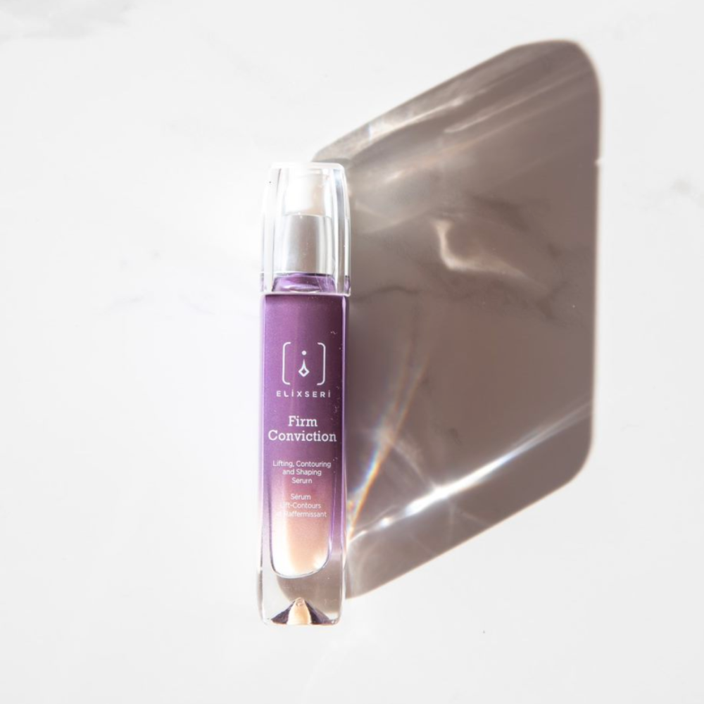 A purple glass bottle of Elixseri Firm Conviction serum on a white marble background with a shadow.