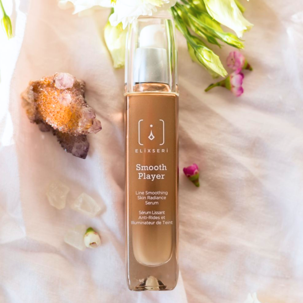 An orange bottle of Elixseri Smooth Player serum amongst flowers and crystals.