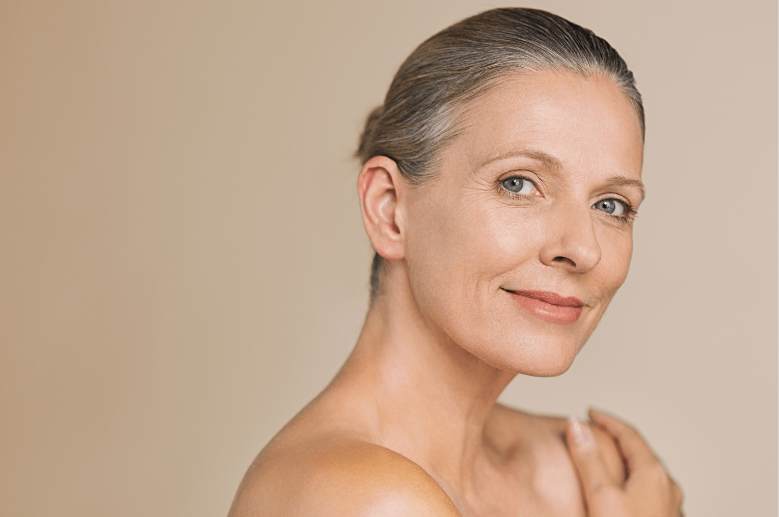 A grey haired middle aged woman with bare skin smiling at the camera. Discussing how to care for your eye and neck area.