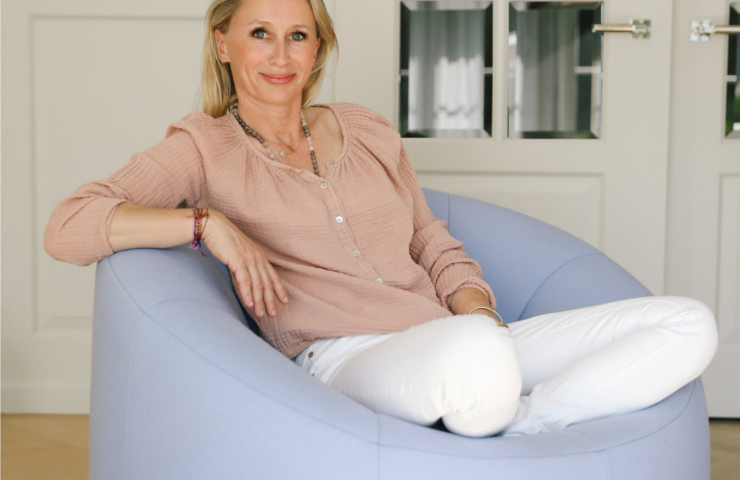 Une femme blonde assise en tailleur sur une chaise ronde bleue dans son salon, souriant en regardant la caméra.