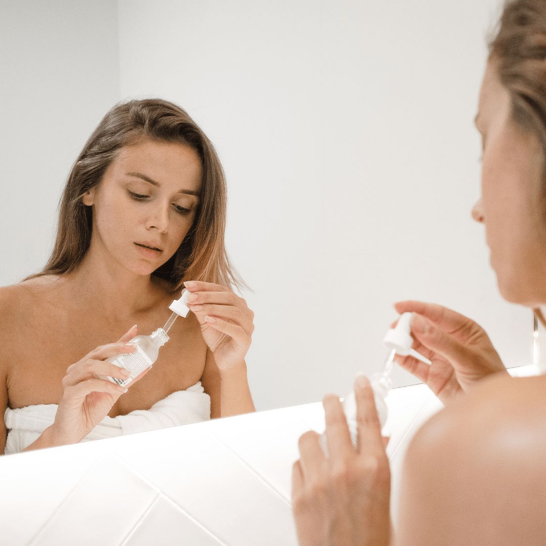 Une femme brune face au miroir dans une salle de bains, portant une serviette et appliquant un sérum pour le visage.