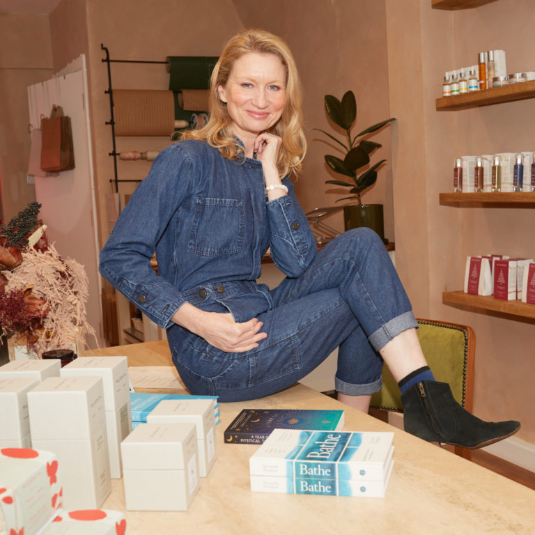 Suzanne Duckett wearing a denim jumpsuit sat on a table in her wellbeing and lifestyle shop, Onolla