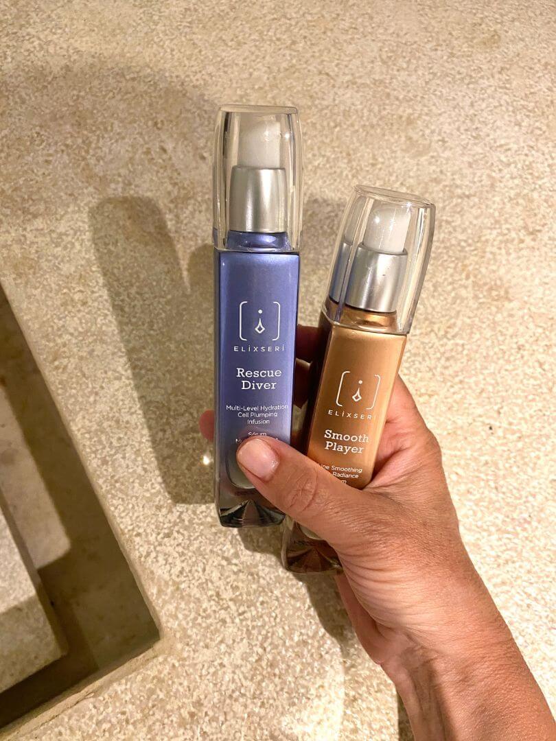 A blue bottle of Rescue Diver and goldy orange bottle of Smooth Player Elixseri serums being held against a beige stone surface. Lighter serums for spring time