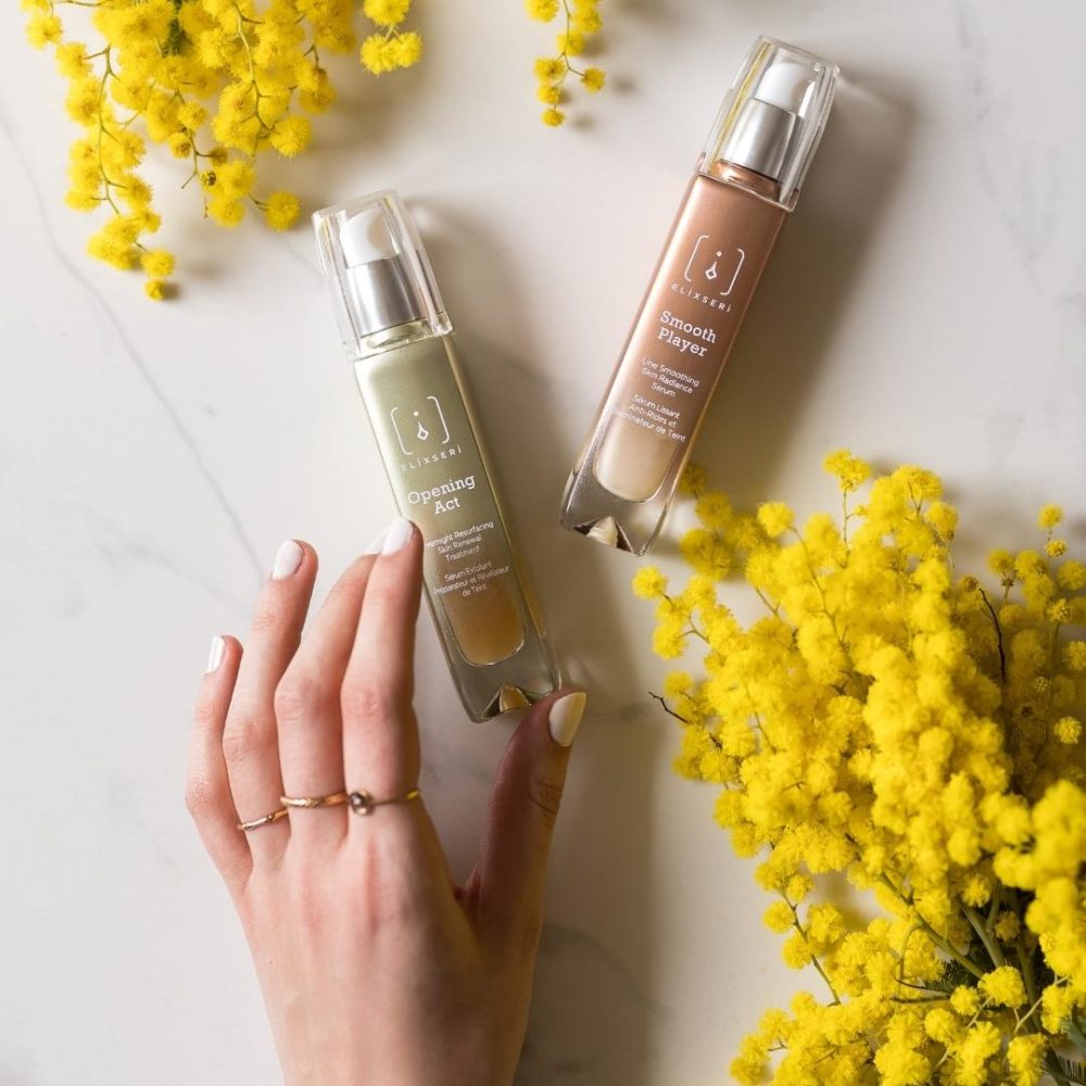 Opening Act and Smooth Player anti-ageing serums pictured with mimosa flowers and a hand with white nail polish
