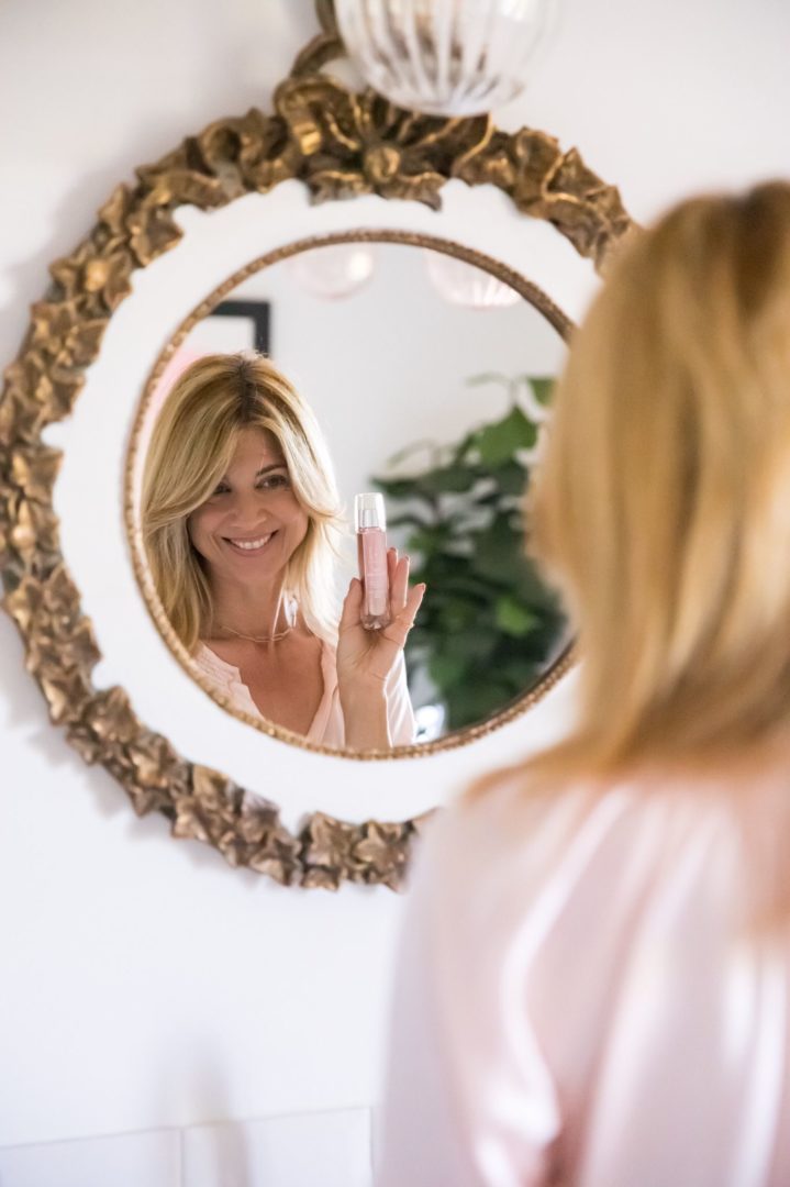 Blonde woman looking at herself in the mirror holding Elixseri Skin Meditation in her hand.