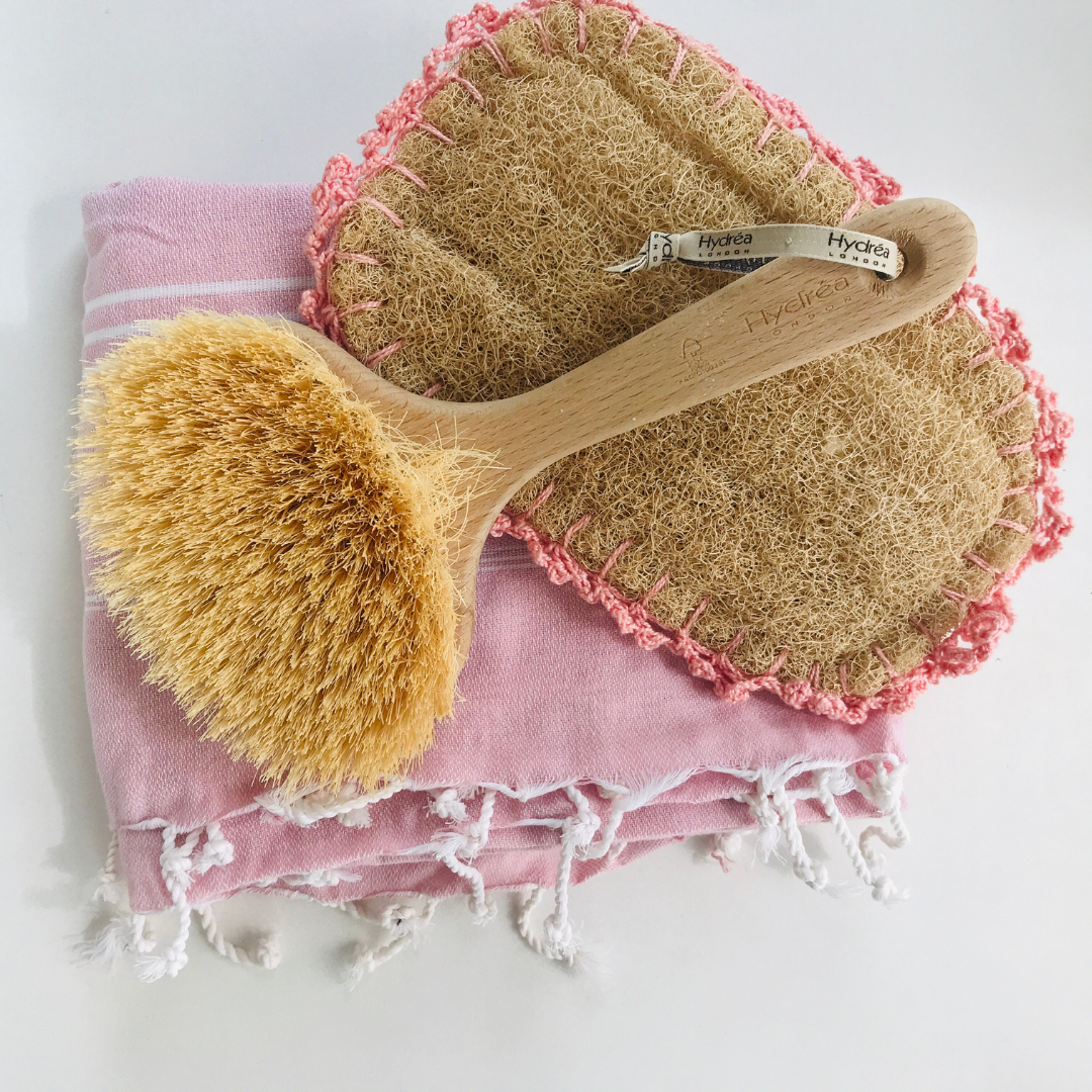 Eine trockene Körperbürste und ein Luffa über einem rosafarbenen Handtuch, um das trockene Bürsten des Körpers als Teil der Förderung einer gesunden Sommerhaut zu diskutieren