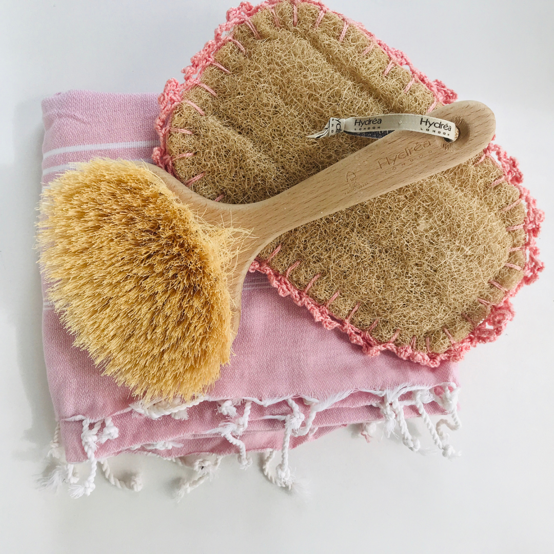 Une brosse pour le corps et un luffa sont présentés sur une serviette rose pour discuter du brossage du corps à sec dans le cadre de la promotion d'une peau estivale saine.