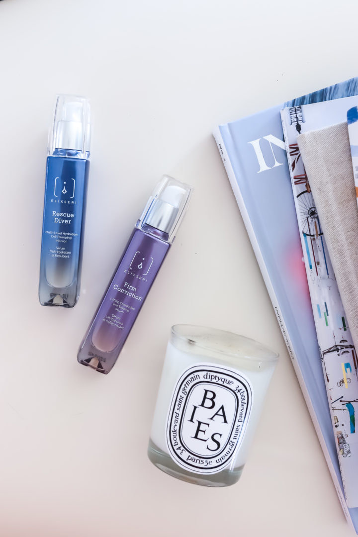 Elixseri's Rescue Diver and Firm Conviction oil free serums pictured with a luxury candle and summer travel magazines
