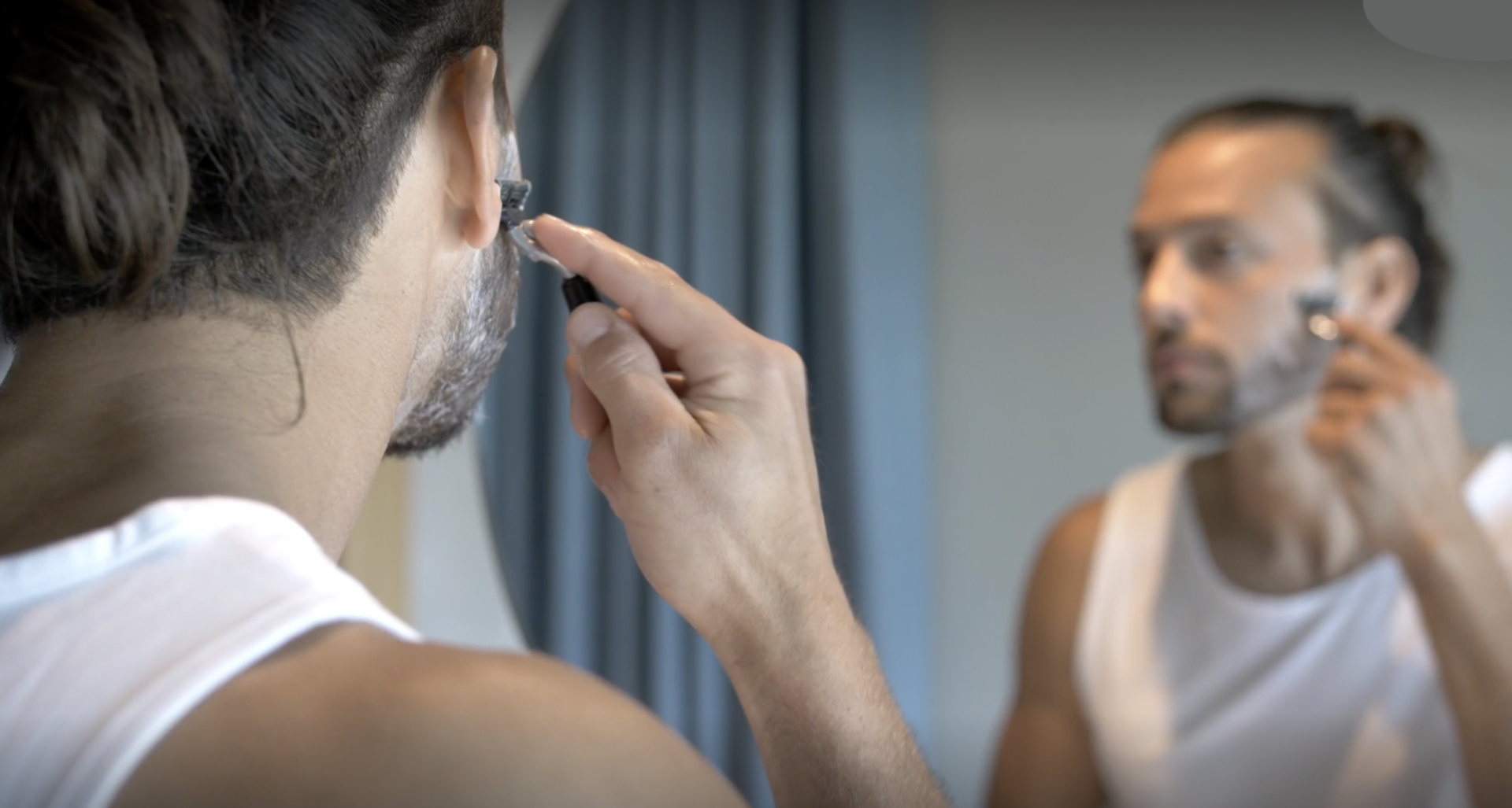 A man shaving in the mirror with a razor as part of his skincare routine