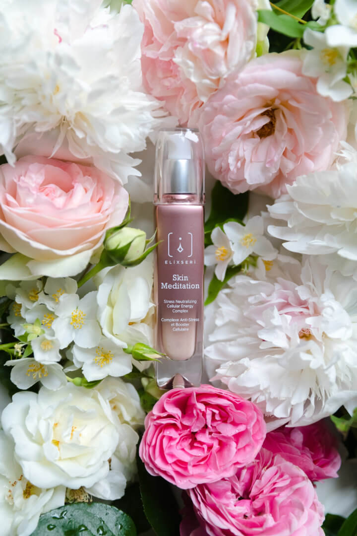 Sérum pour le visage Skin Meditation d'Elixseri, complexe d'énergie cellulaire neutralisant le stress. Le flacon est couché parmi un lit de fleurs roses et blanches. Gagnant des Hip & Healthy 2022 Beauty Awards.