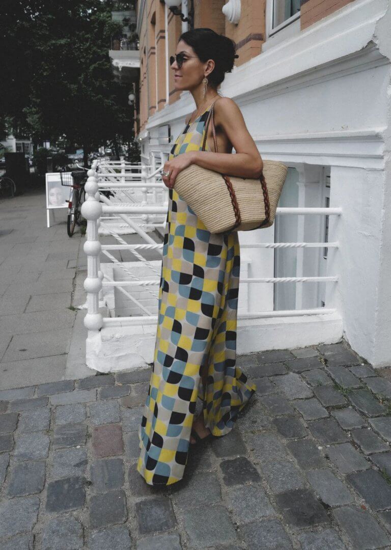 Georgina Moreno wearing sunglasses and a hippie blue and yellow print maxi dress with a straw designer bag. 