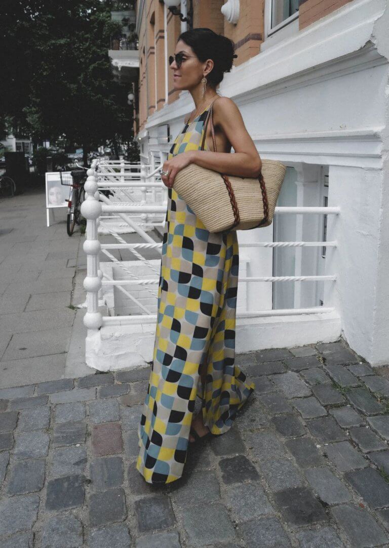 Georgina Moreno porte des lunettes de soleil et une maxi robe hippie à imprimé bleu et jaune avec un sac en paille de créateur. 