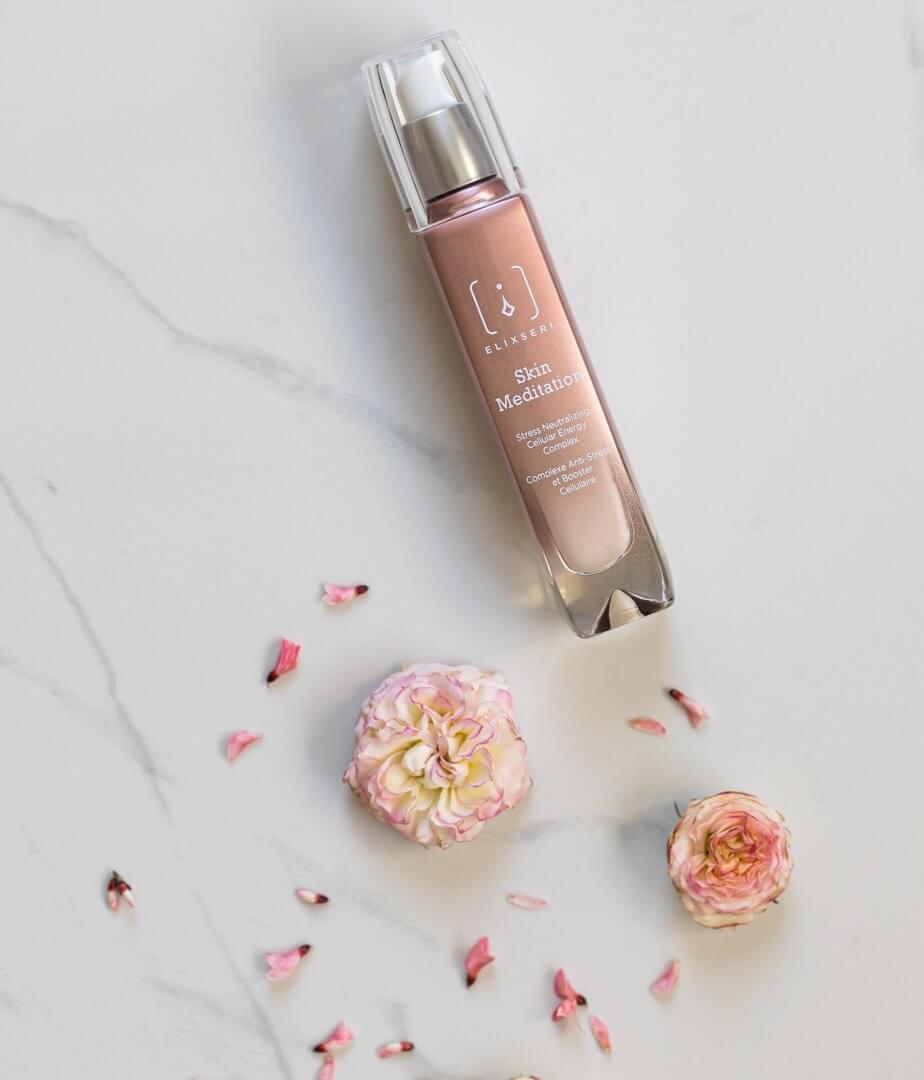 Eine bräunlich-rosa Glasflasche mit dem Serum Elixseri Skin Meditation auf einem weißen Marmorhintergrund, auf dem rosa Blumen verstreut sind.