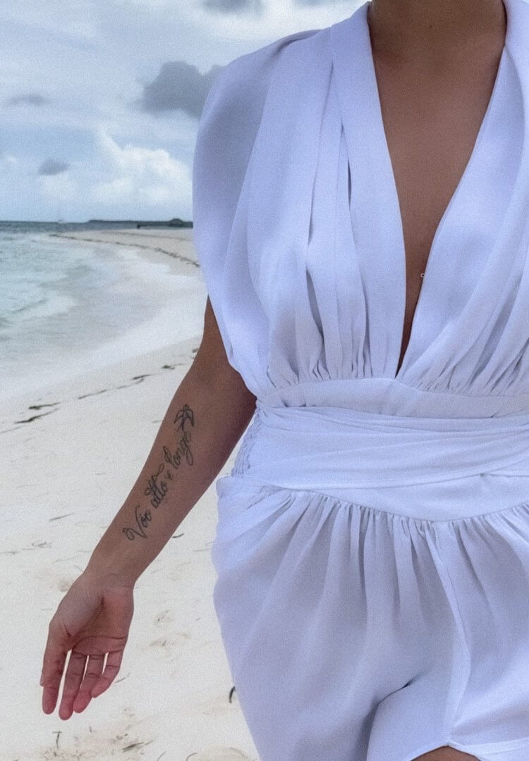 Georgina Moreno wearing a white dress on a beach with her tattooed arm visible. 