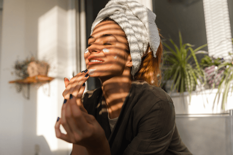 A smiling woman sitting in the sun wearing a white hair towel applying skincare to her face