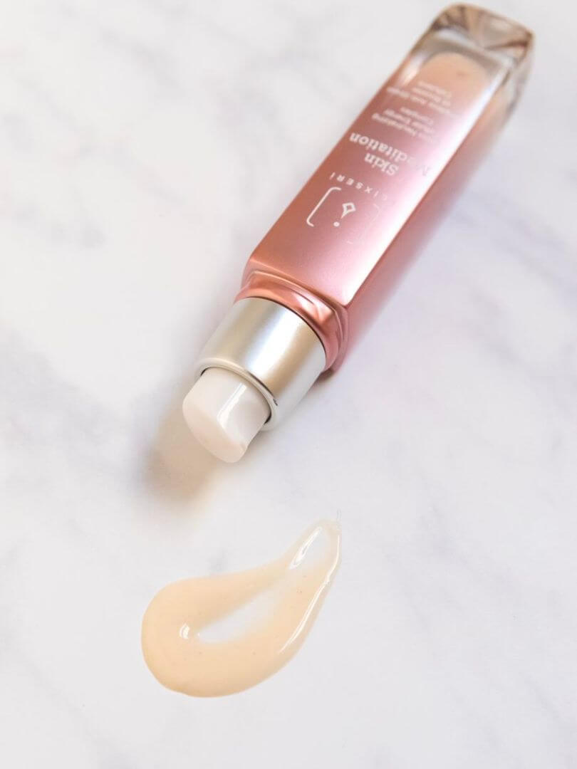 A dusty rose glass bottle of Elixseri Skin Meditation lying on its side against a pale grey marble background with a drop of the creamy serum on the marble.