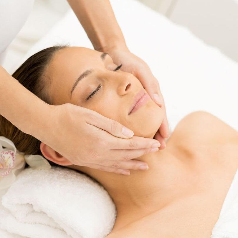 A woman enjoying a relaxing facial with a rolled up towel behind her neck  while the therapist gives her a treatment in a serene room.