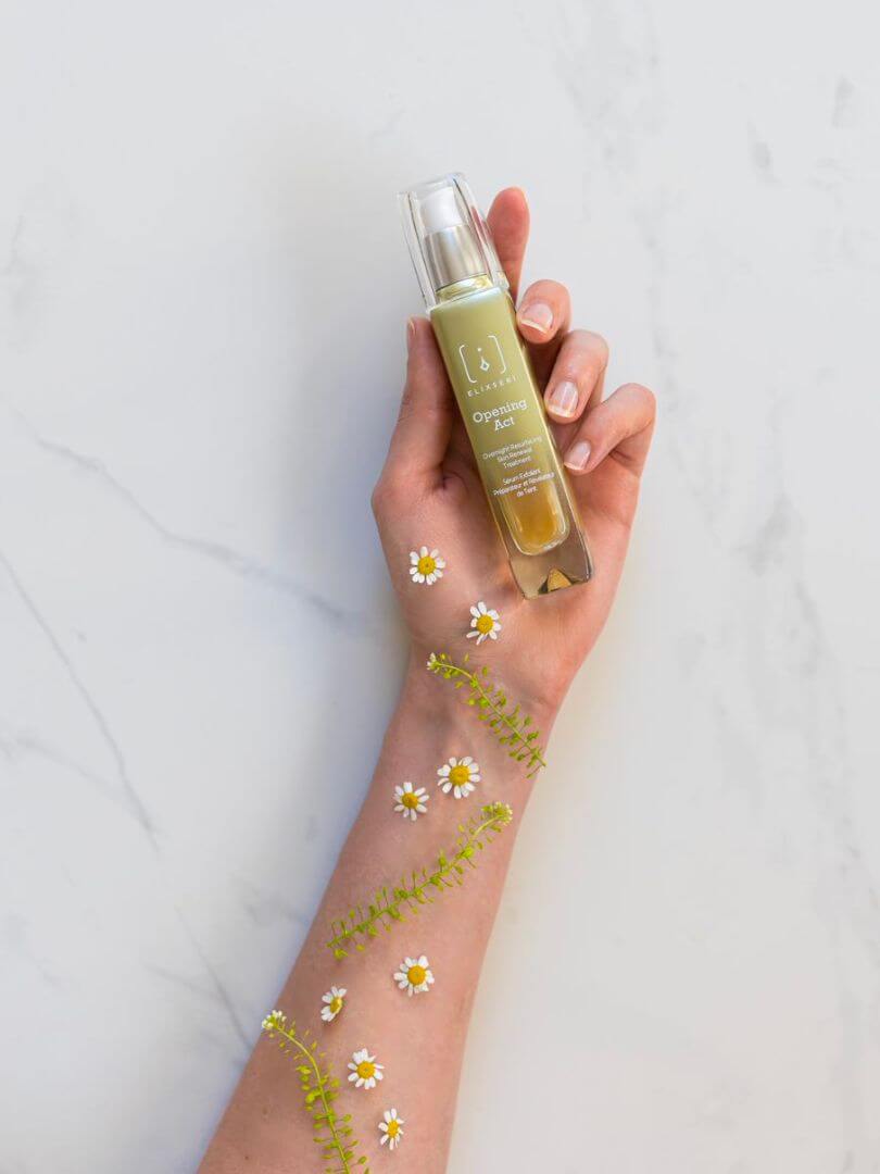 A pale green glass bottle of Opening Act Elixseri serum held in a hand covered in daisies to represent exfoliating spring skincare