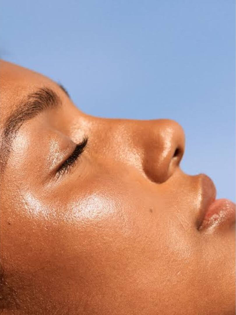 A tanned woman facing the sun with her eyes closed getting sunshine on her face with glowing skin.