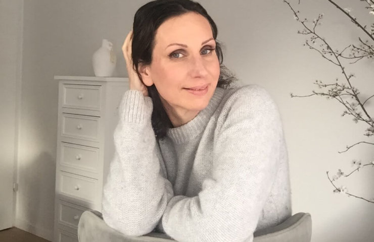 A pretty lady with dark hair and pale skin, sitting on a chair with flowers in the background.