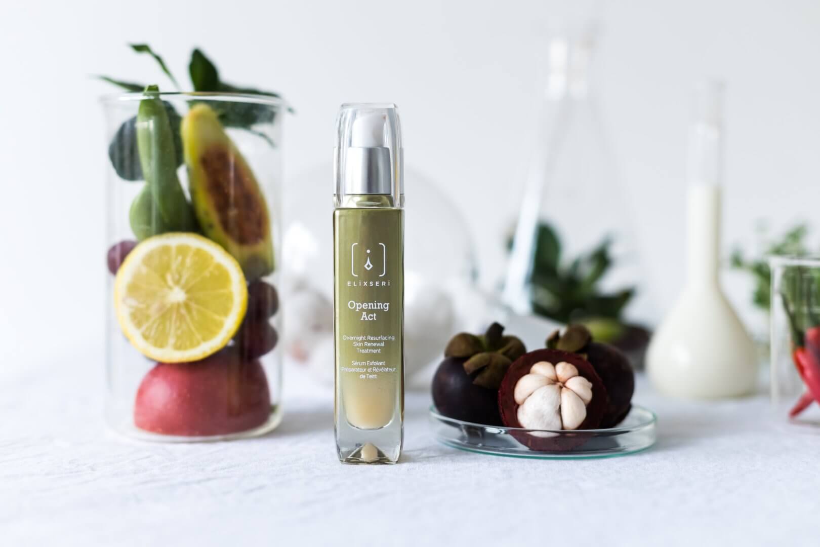 A green bottle of Elixseri Opening Act serum standing amongst AHA fruits and serum ingredients on a white background