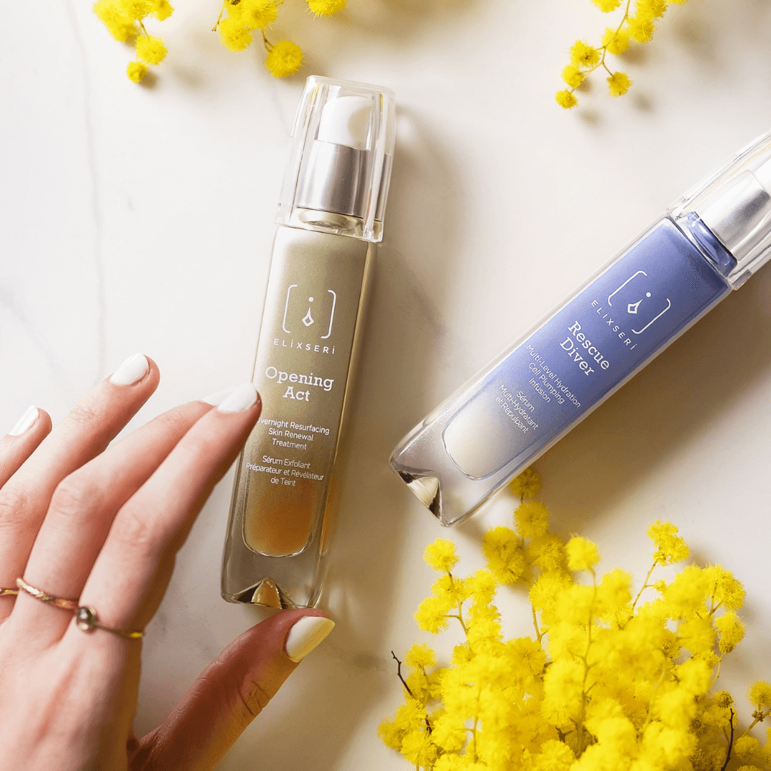 The Elixseri Fresh Start serum duo - a green glass bottle of Opening Act and a blue glass bottle of Rescue Dive on a pale marble background with yellow mimosa flowers and a lady's hand reaching for the Opening Act bottle.