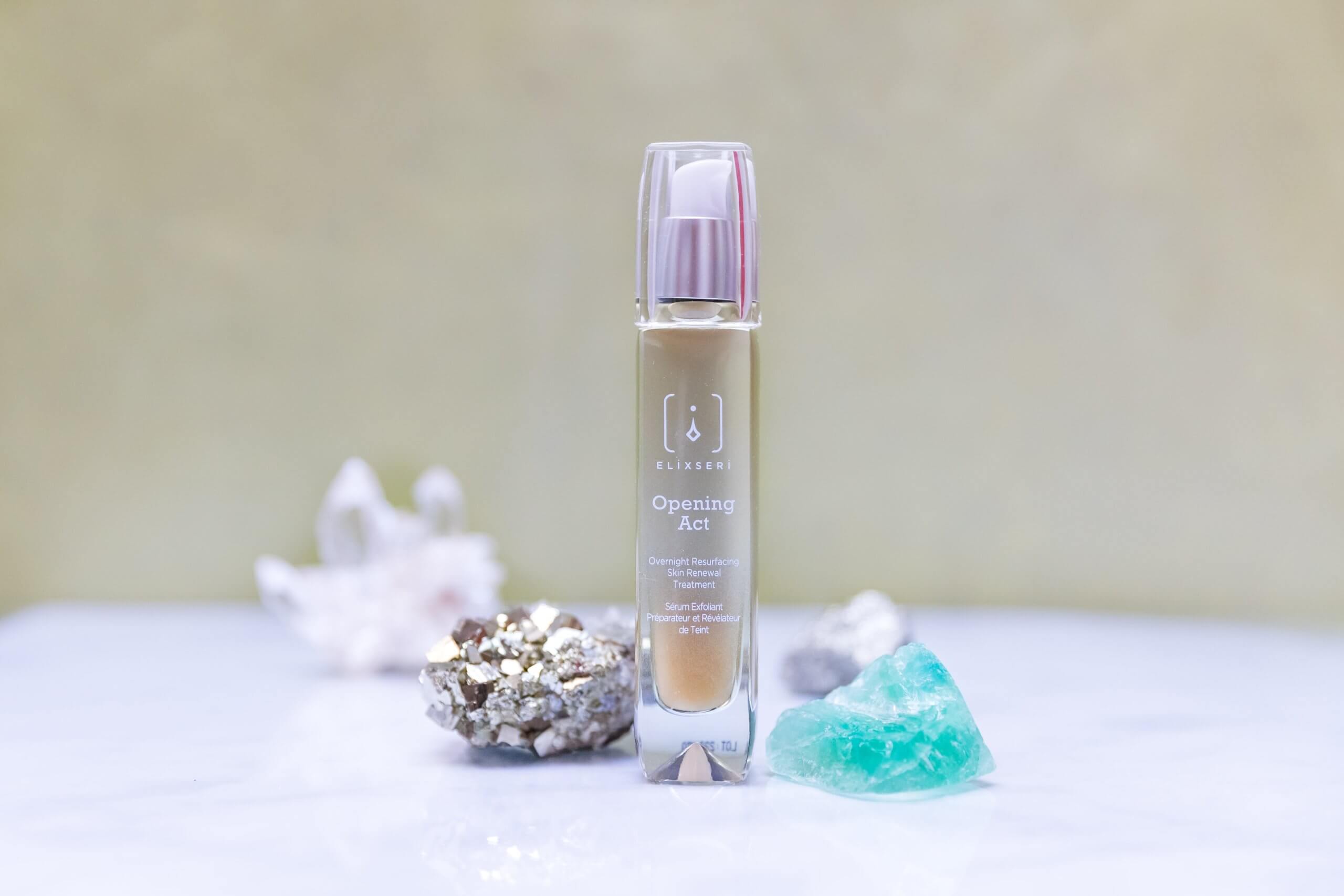 A green bottle of Elixseri Opening Act serum standing on a sandy green background set amongst a collection of crystals