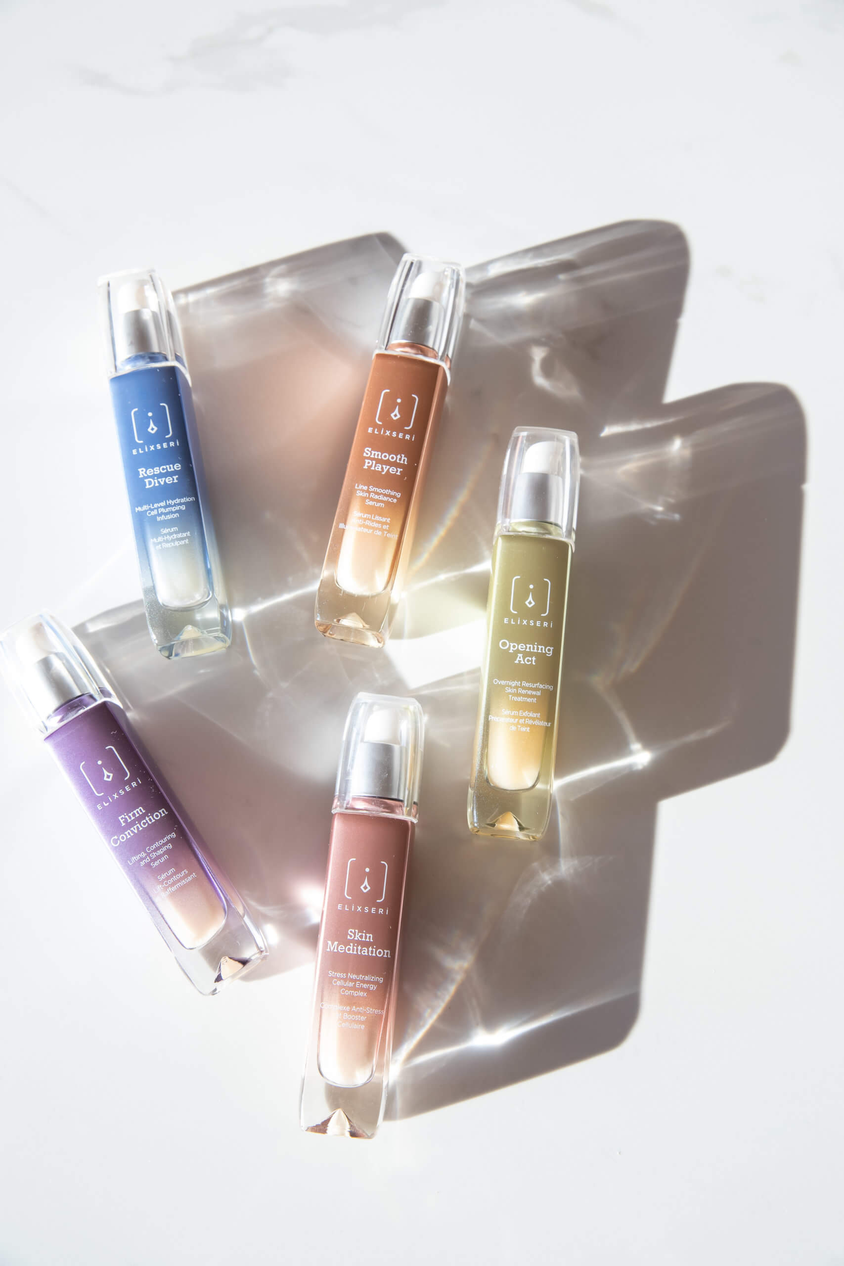 Coloured glass Elixseri serum bottles on a white marble background with light shadows.