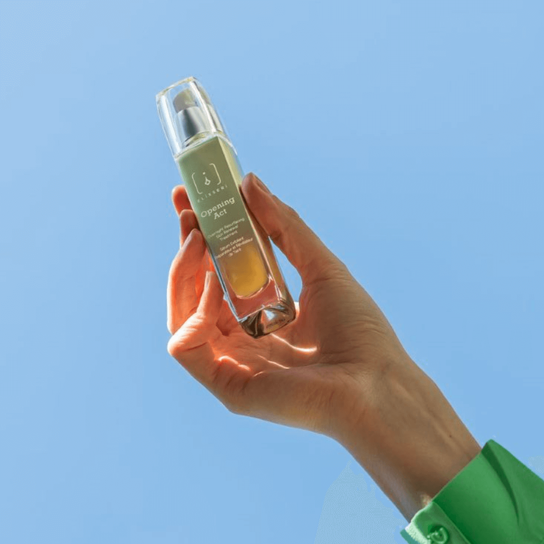 A green glass bottle of Elixseri exfoliating serum 'Opening Act' being held up in the bright blue sky.