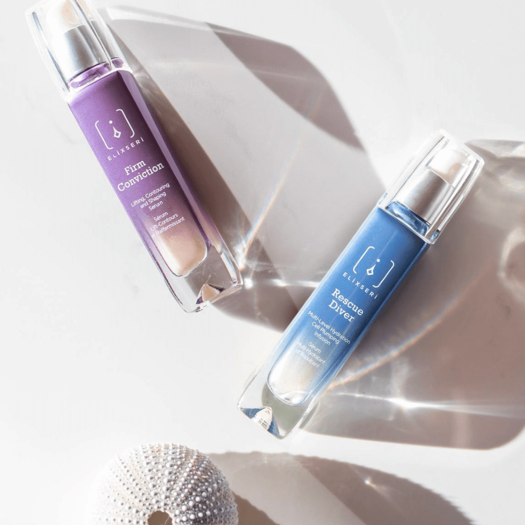 Elixseri's oil free serums, 'Rescue Diver' and 'Firm Conviction' blue and purple glass bottles on a light grey background with a sea urchin shell nearby.