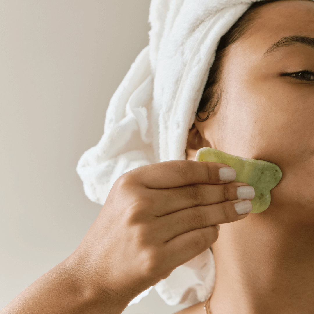 A woman wearing a towel in her hair giving herself gua sha with a jade stone as part of a good summer skincare routine.