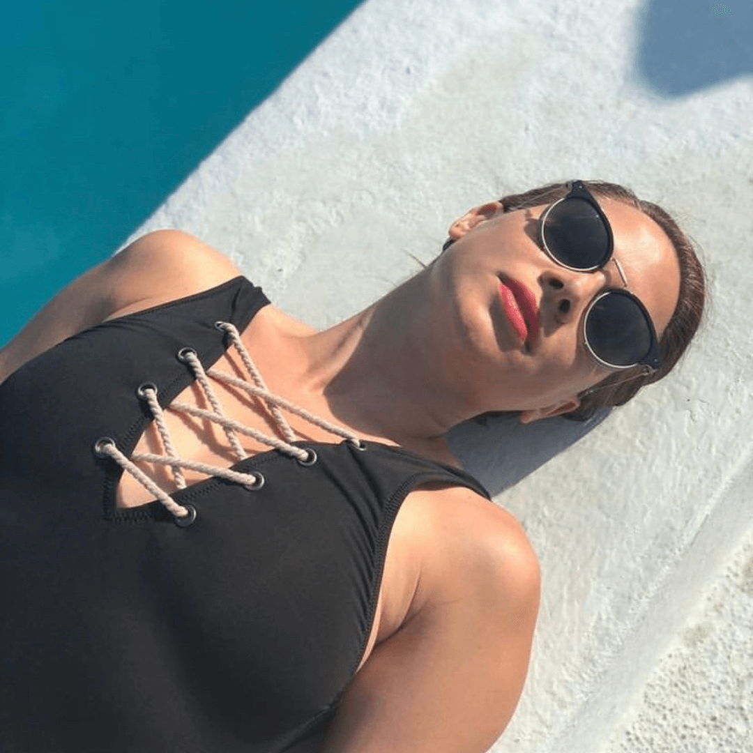 A brunette woman sunbathing wearing a black swimsuit with red lipstick and dark sunglasses. Protecting lips and eyes in summer.