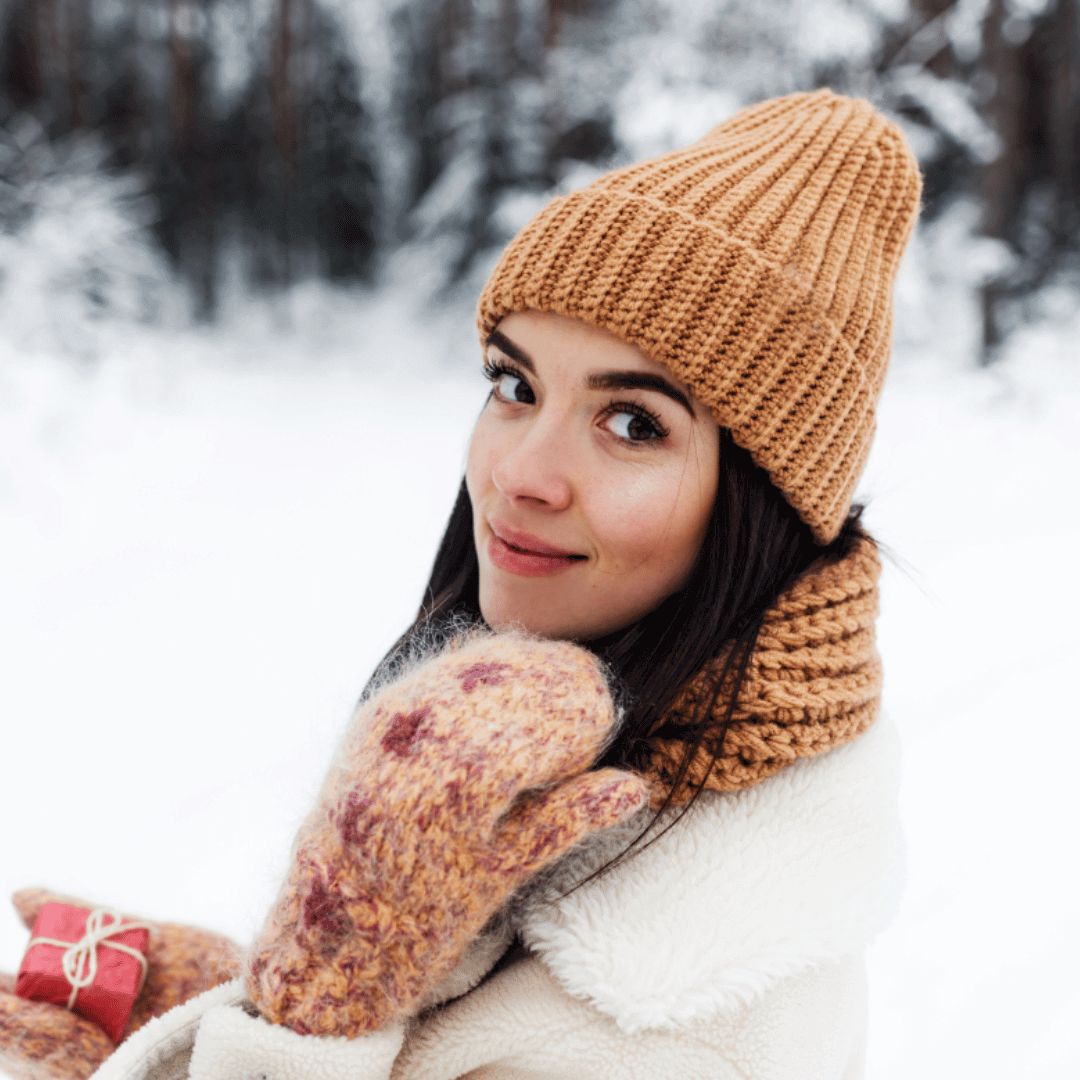 Eine dunkelhaarige Frau in einer verschneiten Umgebung lächelt in die Kamera und trägt Handschuhe und einen Hut.