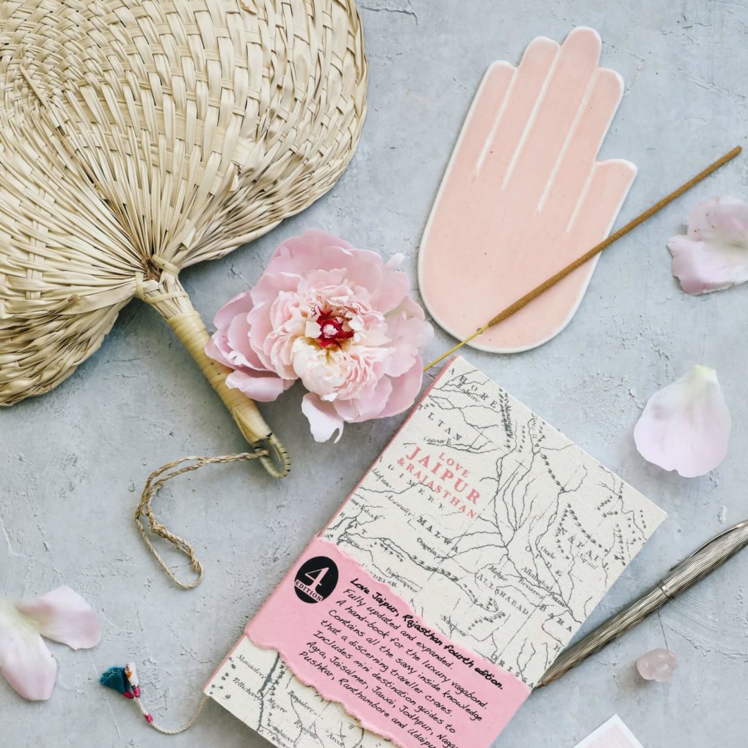 Ein Buch über Rajasthan in Indien, umgeben von rosafarbenen Accessoires und rosafarbenen Blumen auf hellgrauem Hintergrund, daneben ein handgefertigter Fächer aus Seegras, ein Stift und ein Räucherstäbchen.